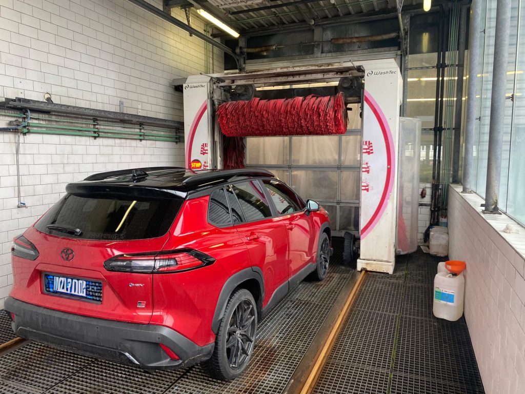 Roter Toyota SUV mit blauem Kennzeichen steht in automatischer Waschanlage mit weißen Fliesenwänden und Gitterboden. Roter Toyota SUV mit blauem Kennzeichen steht in automatischer Waschanlage mit weißen Fliesenwänden und Gitterboden.